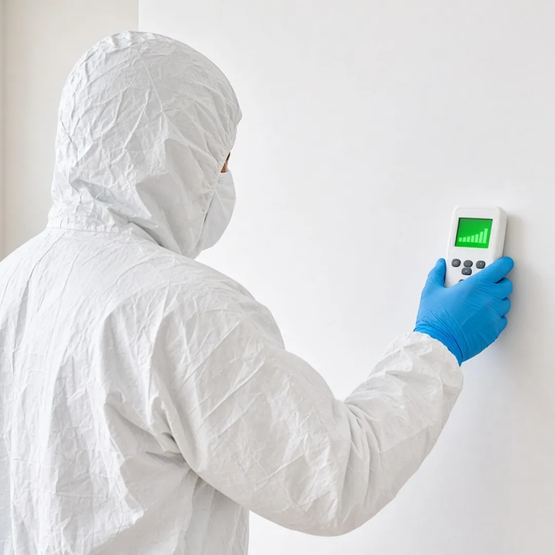 A biohazard remediation technician in a white Tyvek suit and N95 respirator holding an ATP testing meter against a white wall surface in a residential room